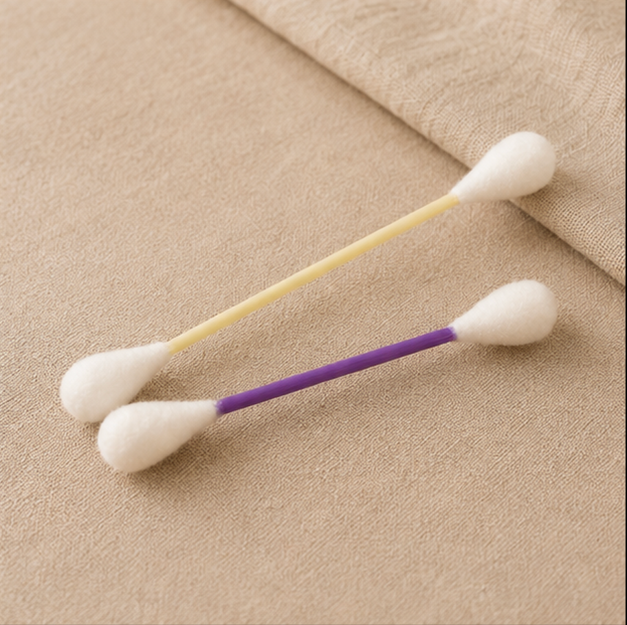 Two cotton swab sticks with white cotton tips and colored plastic handles, one with a pale yellow handle and one with a purple handle, resting on a beige textured fabric background.