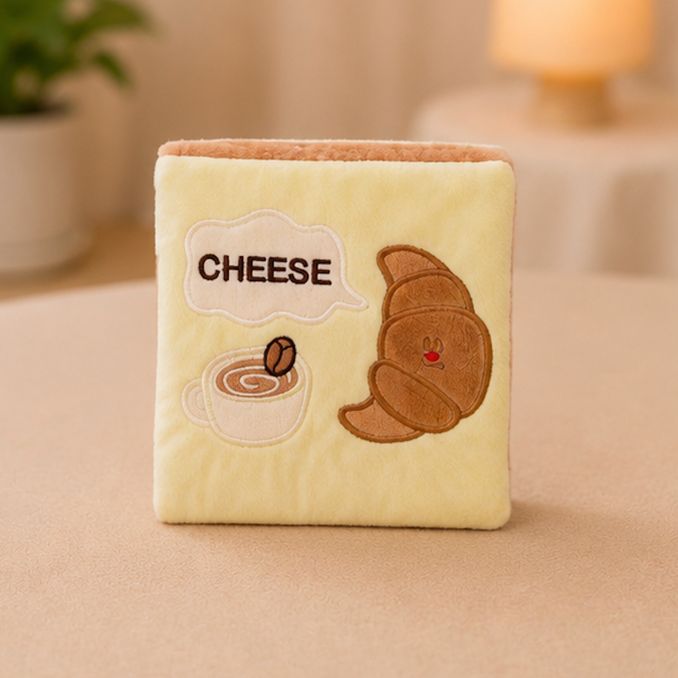 A square cream-colored plush cat snuffle wallet featuring embroidered details of a croissant, a coffee cup, and a speech bubble reading 'CHEESE', shown on a beige tabletop with a softly blurred indoor background.