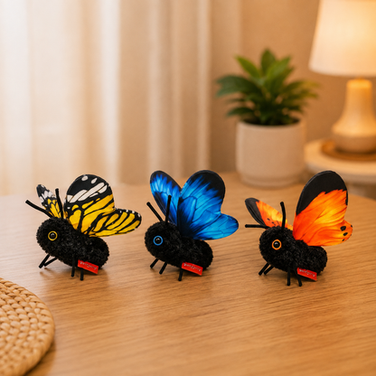 Three black fuzzy toy butterflies with colorful wings are lined up on a wooden table in a softly lit room. The wings show yellow-and-black, blue, and orange patterns, with a small red tag attached near the bottom of each.