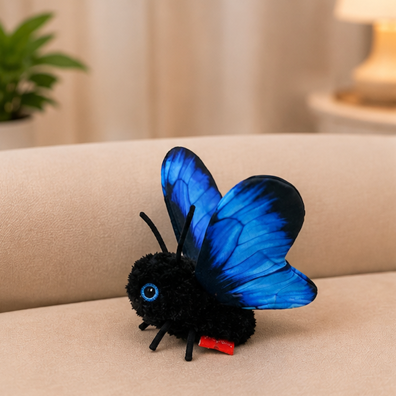 A black plush butterfly toy with vibrant blue wings and a small red tag, resting on a beige fabric surface in a softly blurred indoor setting.