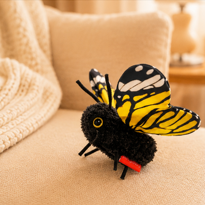 A plush toy Butterfly with black fuzzy body, yellow and black striped wings, and a small red accessory on its leg, resting on a beige fabric surface with a soft home background.