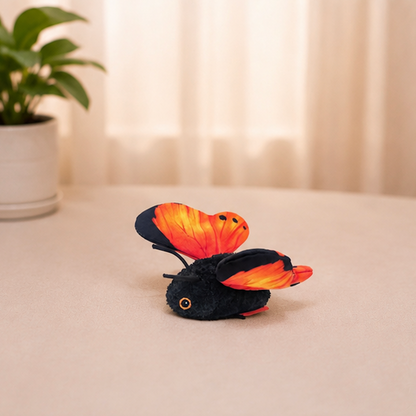 A small plush toy butterfly with black fuzzy body and bright orange-red wings resting on a beige tabletop in a softly lit indoor setting with a blurred plant in the background.