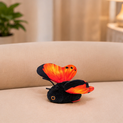 A black plush toy butterfly with bright orange and red wings resting on a beige fabric surface in a softly blurred indoor setting.