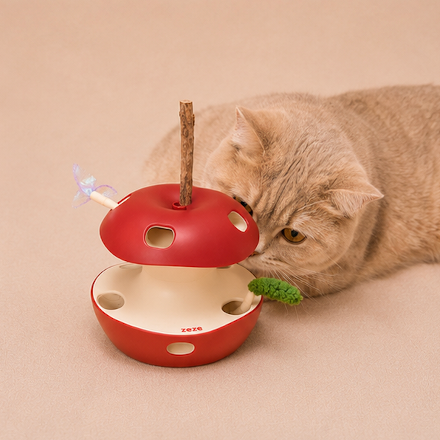 A beige cat lies on a warm beige surface, interacting with a red apple-shaped cat toy that has a wooden stick inside and a small green textured piece attached.