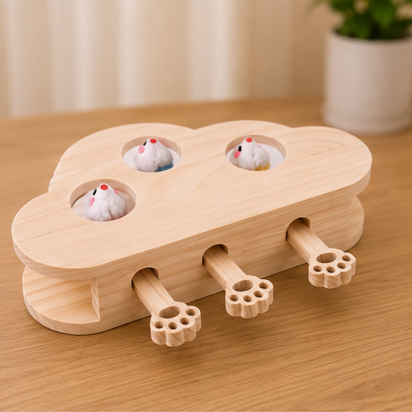 A wooden cloud-shaped toy or puzzle with three paw-shaped handles and small hamster holes on top, shown on a wooden table with hamsters peeking out. A blurred plant and curtain appear in the background.