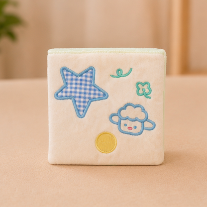 A square cream-colored plush cat snuffle wallet with embroidered cartoon motifs, including a blue-and-white gingham star, a blue sheep face, a green four-leaf clover, and a yellow circle, shown on a soft beige surface.