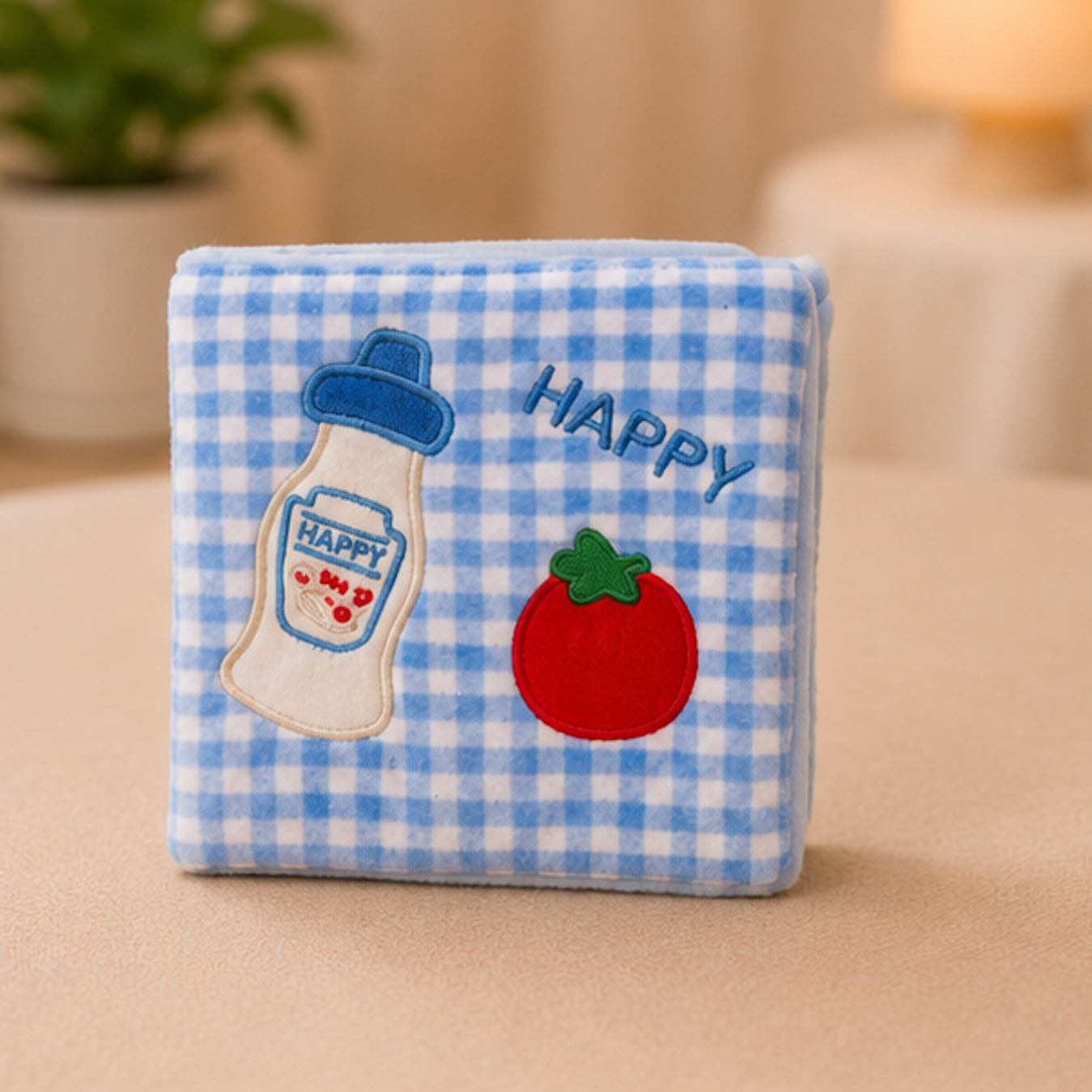 A square plush cat snuffle wallet featuring a blue and white gingham fabric pattern, embroidered with a milk bottle, a red tomato, and the word 'HAPPY' on a soft beige surface.