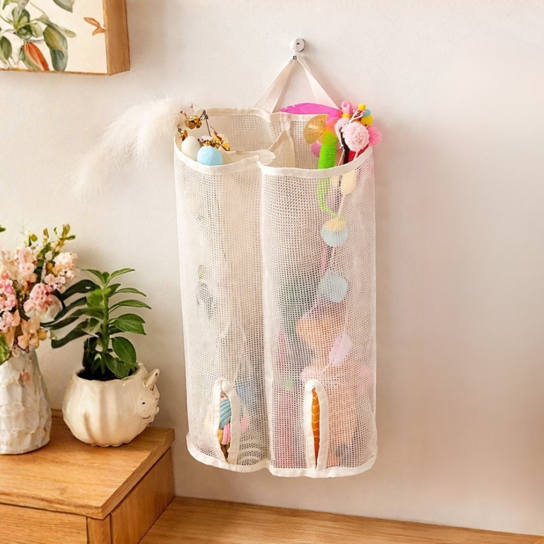 Hanging mesh storage bag with cat toys on a wall next to a wooden shelf with plants and a picture frame.
