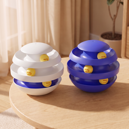 Two small spherical toy-like products with concentric ring layers are displayed on a round wooden table in a softly lit indoor setting. One is white with blue accents and yellow smiley-face details, and the other is blue with white accents and matching yellow smiley details.