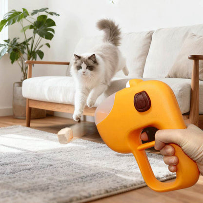 A fluffy white Ragdoll cat jumping off a sofa to chase a treat launched from an orange handheld treat dispenser in a bright living room.
