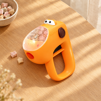 An orange handheld cat treat launcher with a clear dome filled with treats, resting on a wooden table in sunlight next to a small bowl of treats.