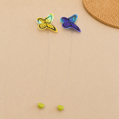 Two replacement butterfly attachments—one yellow and one purple—mounted on flexible wires with green bases, sitting on a wooden surface in a brightly lit room.