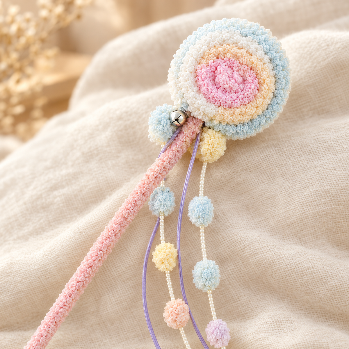 Close-up of a plush pastel swirl cat teaser wand head with a tiny bell and colorful pom-pom strands, resting on a beige cloth.
