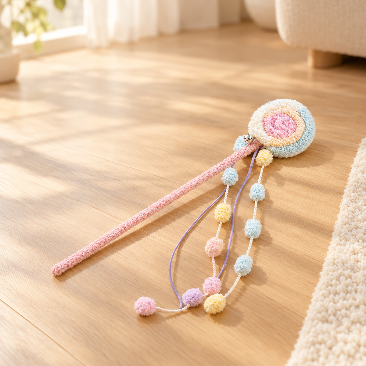 A plush pastel swirl cat teaser wand with a pink handle, round lollipop top, and colorful pom-pom strands, laid out flat on a light wood floor next to a cream rug.