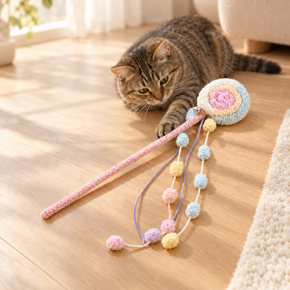 A brown tabby cat lying on a light wood floor, reaching its paw toward a plush pink swirl cat teaser wand with pastel pom-pom strands.